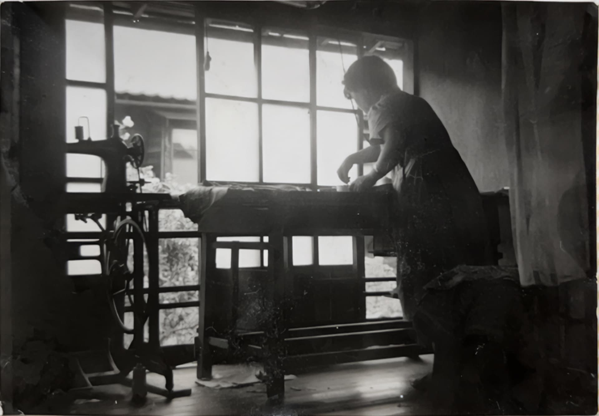 白黒写真。窓際で女性が服を仕立てている